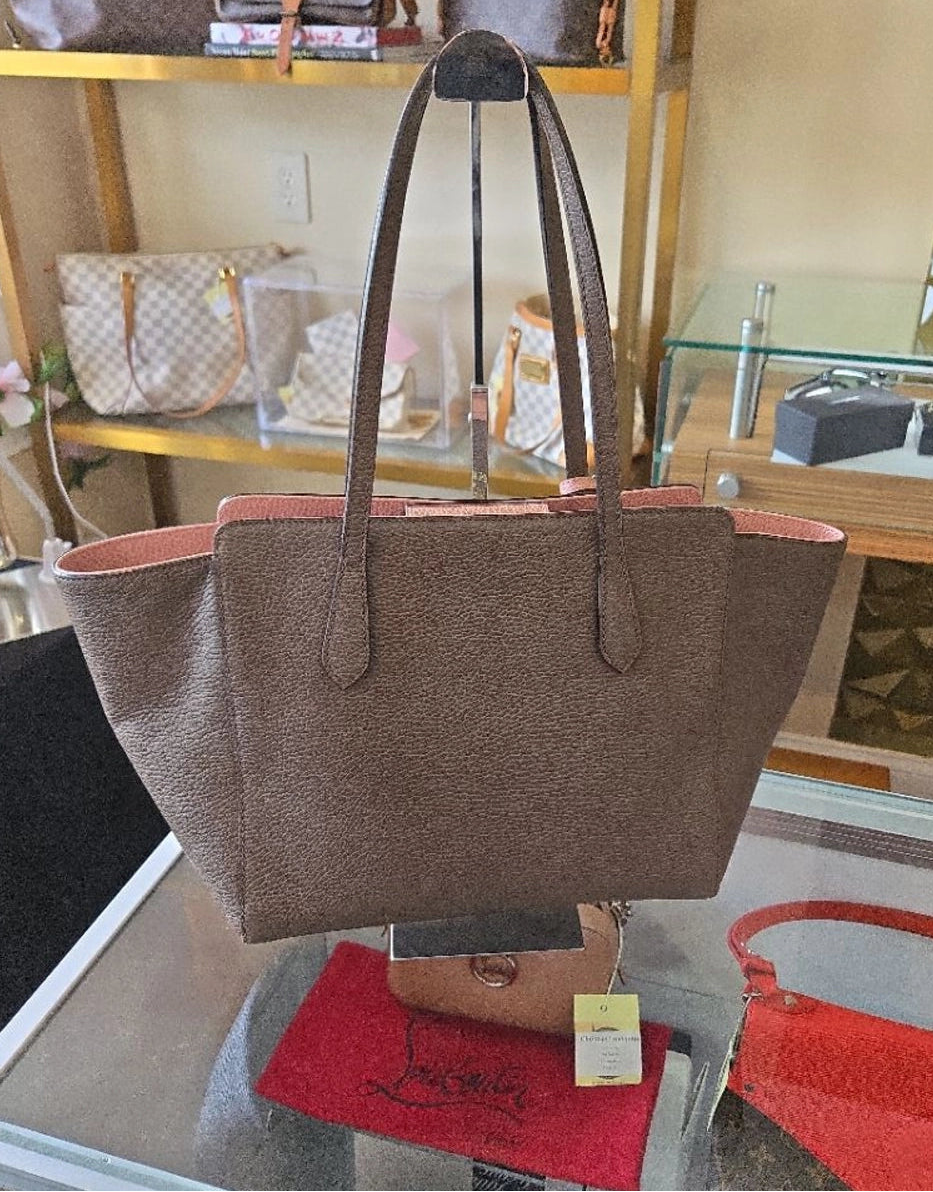 A brown textured leather tote bag with pink lining and gold-tone hardware, displayed on a glass table inside a retail store.