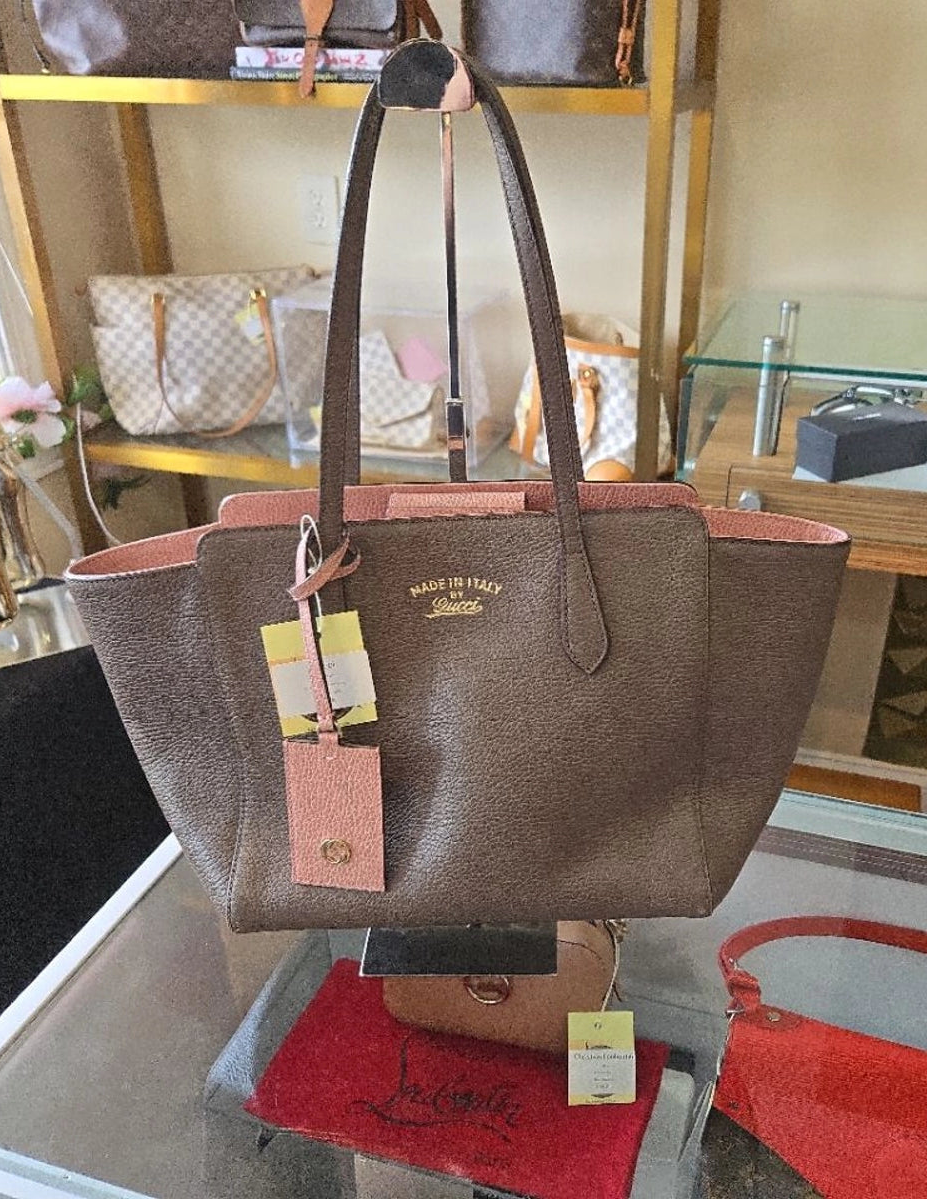 A brown textured leather tote bag with pink lining and gold-tone hardware, displayed on a glass table inside a retail store.